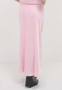 Jupe maxi en satin rose à la silhouette fluide, avec une texture douce et un léger éclat, associée à des bottines noires à la cheville.