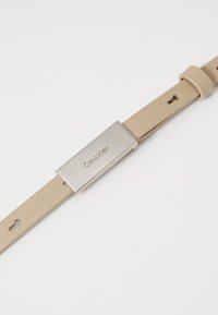 Bež remen od kože za sat s kopčom od srebrnog nehrđajućeg čelika, sa logotipom "Calvin Klein" i više rupa za podešavanje.