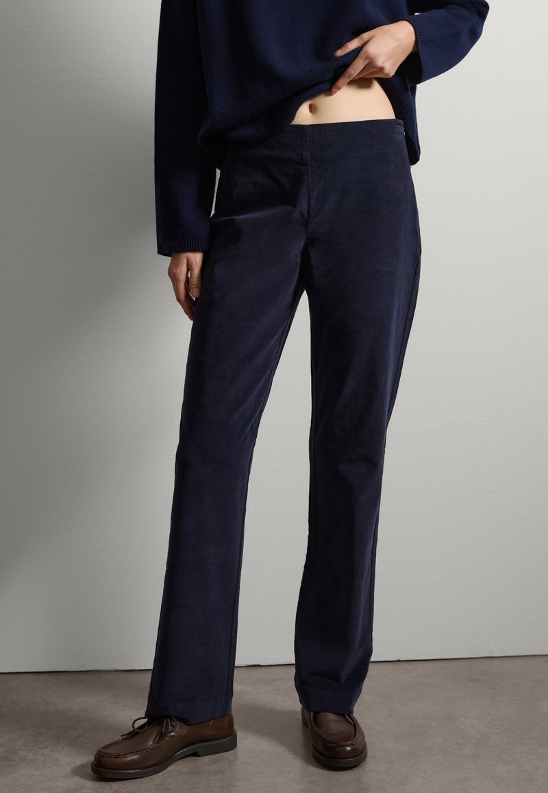 Pantalons en velours côtelé bleu marine avec une coupe droite, présentant une texture lisse et un éclat subtil, associés à des chaussures en cuir marron.
