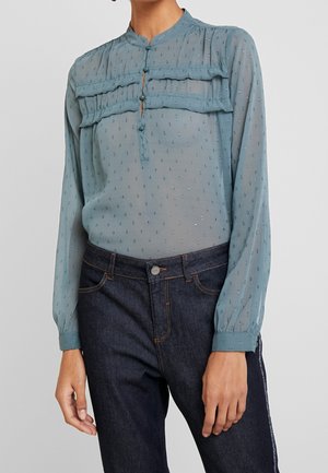 Femme portant un chemisier léger bleu clair transparent à petit motif à pois et détails volantés, associé à un jean bleu foncé.