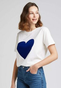 T-shirt en coton blanc avec un grand motif de cœur bleu marine sur le devant. Manches courtes, col rond, associé à un jean bleu.