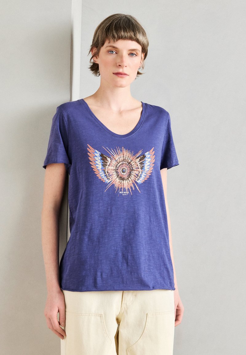 Μπλουζάκι κοντομάνικο με V-λαιμό σε μωβ ύφασμα, που διαθέτει μια πολύχρωμη γραφιστική σχέδιο με φτερά και κεντρικό κυκλικό μοτίβο. Μαλακή υφή.