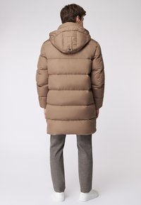 Giacca a vento in marrone chiaro, con cappuccio, design trapuntato e lunghezza a metà coscia. Indossata sopra pantaloni grigi e scarpe bianche.
