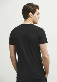 Mężczyzna z brązowymi włosami, ubrany w prostą czarną koszulkę z krótkim rękawem, stoi tyłem do aparatu, z głową obróconą w prawo.