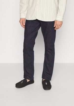 Homme portant un pantalon bleu foncé à jambes droites, des mocassins noirs avec un logo blanc, et une chemise claire rayée rentrée, debout sur un sol uni.