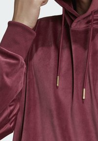 Samettinen viininpunainen huppari, jossa on kiristysnauhallinen huppu, resorireunaiset hihansuut ja kullanväriset metalliset nauhanpäät. Pehmeä rakenne, jossa sileä ja kiiltävä pinta.