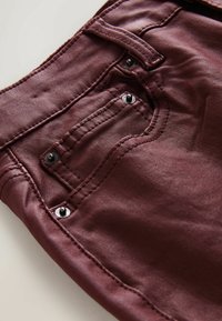 Los jeans de mezclilla burdeos cuentan con un bolsillo trasero con remaches negros y detalles de costura sutiles. La tela tiene una textura suave y lisa.