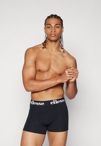 Schwarze Boxerbriefs mit glatter Textur und elastischem Bund, der ein weißes Logo trägt. Das Modell hat geflochtenes Haar und ist oberkörperfrei.