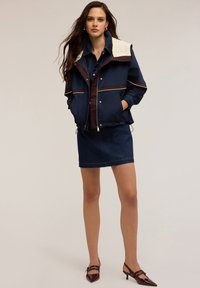 Giubbotto leggero blu navy con colletto crema e dettagli marroni, abbinato a una gonna di jeans e scarpe con tacco marroni.
