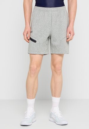 Mann trägt hellgraue Sportshorts mit seitlicher Reißverschlusstasche, weiße Socken und weiße Sportschuhe, steht vor schlichten Hintergrund.