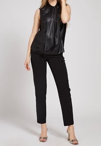 Blouse noire sans manches avec un col noué et un motif de petits pois subtils, accompagnée d'un pantalon noir slim et de talons ouverts à motifs.