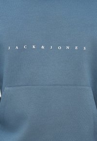 Μπλε φούτερ με μπροστινή τσέπη, στρογγυλή λαιμόκοψη και λευκή επιγραφή «JACK & JONES» στο στήθος. Απαλή ύφανση με λεία υφή.