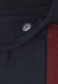 Marineblaues Gewebe mit Rippstruktur, das mit einem klaren Knopfdetail versehen ist. Es gibt einen roten Akzent, der ein Color-Block-Design an der Seite präsentiert.