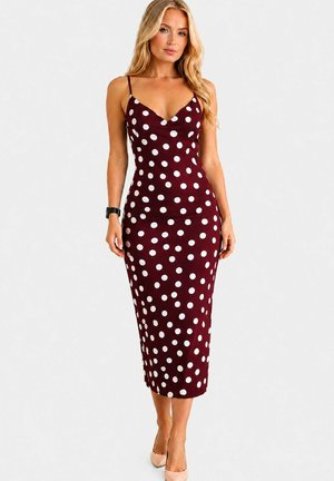 Femme blonde portant une robe ajustée sans manches bordeaux avec des pois blancs et des talons hauts couleur chair, debout devant un fond uni.