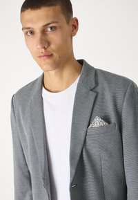 Grå blazer med en strukturerad väv, enfaldig bröstficka som visar en mönstrad näsduk, lager på en vit t-shirt.