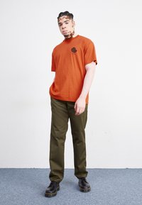 Camiseta naranja de hombre con un logo negro, combinada con pantalones de color oliva y botas negras de cordones. Fondo neutro con alfombra azul.