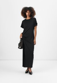 Black maxi dress with short sleeves and a relaxed fit. Smooth texture, featuring a side knot detail. Paired with a small black handbag.