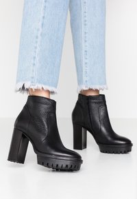 Botas negras hasta el tobillo con parte superior de cuero texturizado, tacón cuadrado y suela serrada, combinadas con jeans de dobladillo deshilachado en azul claro.