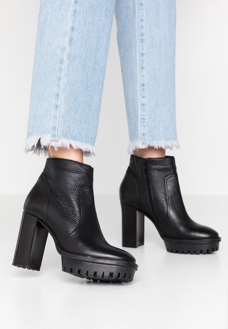 Botas negras hasta el tobillo con parte superior de cuero texturizado, tacón cuadrado y suela serrada, combinadas con jeans de dobladillo deshilachado en azul claro.