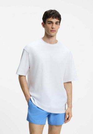 Junger Mann, der ein lockeres weißes T-Shirt und hellblaue Shorts trägt, steht mit einer Hand in der Tasche vor einem schlichten weißen Hintergrund.