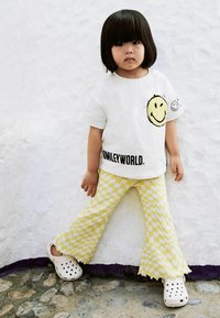 Witte t-shirt met zwarte tekst "SMILEYWORLD" en een gele glimlachende emoji. Gele geruite flared broek. Witte crocs met gaten voor ventilatie.