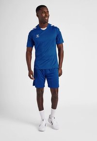 Blaues Sport-T-Shirt mit kurzen Ärmeln und kontrastierenden weißen Streifen an den Schultern, kombiniert mit blauen Shorts und weißen Sportschuhen.