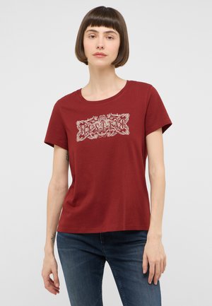 Frau mit kurzen braunen Haaren, die ein rotes Mustang-T-Shirt und blaue Jeans trägt und vor einem schlichten hellen Hintergrund steht.