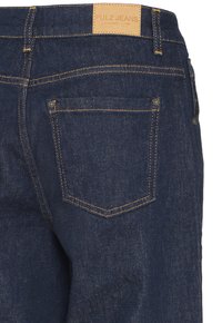 Jean en denim bleu foncé vue arrière montrant la poche arrière droite, coutures jaunes, passants de ceinture et patch en cuir beige avec le nom de la marque Pulz Jeans.
