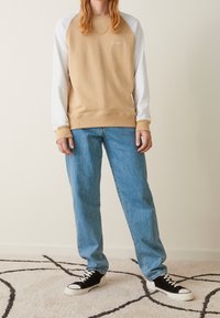 Sudadera beige con mangas raglán blancas, material de algodón, cuello redondo, combinada con jeans azul holgados y zapatillas negras con suelas blancas.