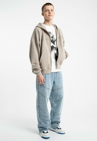 Beżowa bluza z kapturem z przednimi kieszeniami, połączona z luźnymi, jasnoniebieskimi dżinsami z kontrastującymi przeszyciami. Czarno-biała koszulka graficzna pod spodem, oraz sportowe buty z akcentami bieli, szarości i niebieskiego.