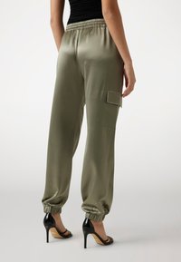 Joggers in satin verde oliva con cintura elastica, polsini alle caviglie e una tasca laterale, abbinati a sandali neri con tacco alto.