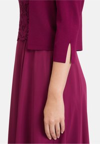 Robe de maternité en magenta profond avec un corsage en dentelle ajusté et une jupe fluide. La manche possède une fente latérale pour faciliter les mouvements.
