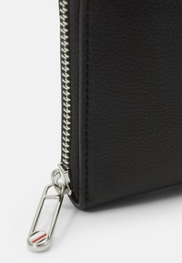 Portafoglio in pelle nera con superficie testurizzata, dettagli in argento sulla cerniera e attacco per portachiavi. Presenta un accento decorativo in metallo.