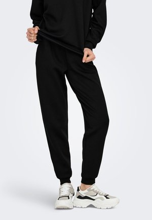 Persona con pantalones de chándal y sudadera negra sujetando el dobladillo de la camisa, de pie con zapatillas gruesas blancas y negras sobre un fondo neutro.