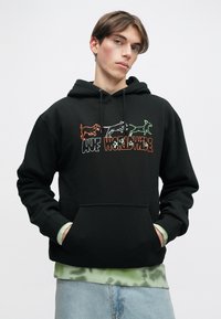 Melna kapuce ar auklu, kurā ir krāsains suņu grafiks un uzraksts "HUFF WORLDWIDE." Novietota virs zaļa taidu krekla.