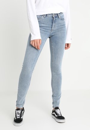 Personne portant un jean skinny bleu clair, un haut blanc à manches longues et des baskets noir et blanc, debout devant un fond blanc uni.