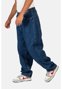 Blue denim jeans with a loose fit, featuring side and back pockets. Paired with white and beige sneakers with red accents.