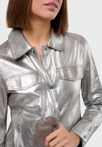 Veste métallique argentée avec un col, deux poches avant, fermeture à pression et manches longues. Texture lisse et brillante.