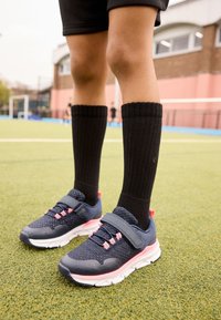 Scarpe sportive blu navy con accenti rosa e una fascia in Velcro, indossate con calzini neri fino al ginocchio, in piedi su erba artificiale.