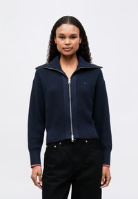 Námornícky úpletový cardigan na zips s vysokým golierom, s strieborným zipsom a červenými akcentmi na manžetách. Nosený s čiernymi nohavicami.