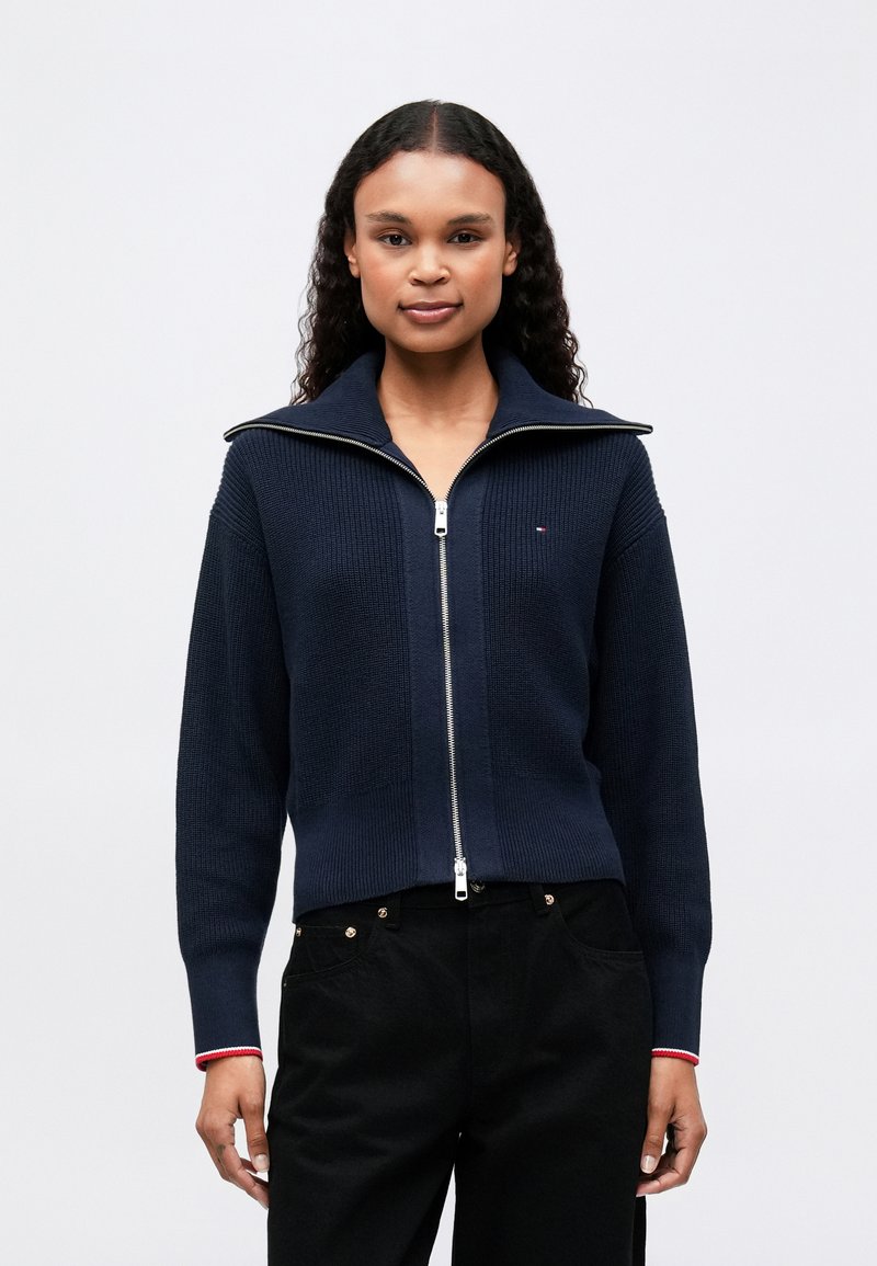 Námornícky úpletový cardigan na zips s vysokým golierom, s strieborným zipsom a červenými akcentmi na manžetách. Nosený s čiernymi nohavicami.