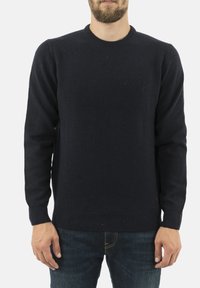 Mężczyzna w prostym czarnym swetrze dzianinowym i ciemnoniebieskich dżinsach, stojący z rękami swobodnie opuszczonymi wzdłuż ciała na białym tle.