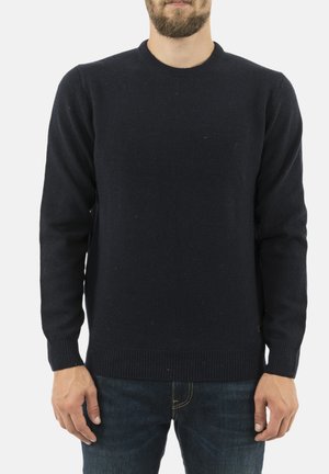 Mężczyzna w prostym czarnym swetrze dzianinowym i ciemnoniebieskich dżinsach, stojący z rękami swobodnie opuszczonymi wzdłuż ciała na białym tle.