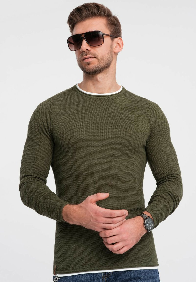 Groene lange mouwen gebreide shirt met een gestructureerd oppervlak en witte bies bij de hals, gecombineerd met een zonnebril en een horloge.