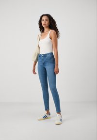Slim-fit, high-waisted blue jeans, paired with a white ribbed tank top and cream shoulder bag. White sneakers with blue stripes.
