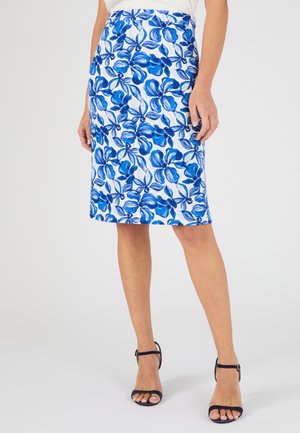 Femme portant une jupe blanche mi-longue à imprimé floral bleu vif, associée à un haut blanc et des sandales à talons hauts bleu marine.