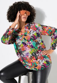 Buntes Langarmbluse mit einem lebhaften Blumen- und Tiergrafikmuster. Kombiniert mit schwarzen figurbetonten Leggings. Das Model trägt große Sonnenbrillen.