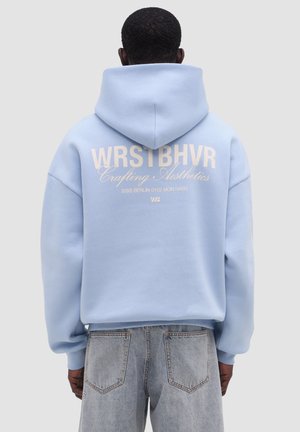 Uomo che indossa una felpa con cappuccio azzurro con la scritta "WRSTBHVR Crafting Aesthetics (030) Berlin 0102 Mon Habit" sulla schiena, abbinata a jeans di denim chiaro.