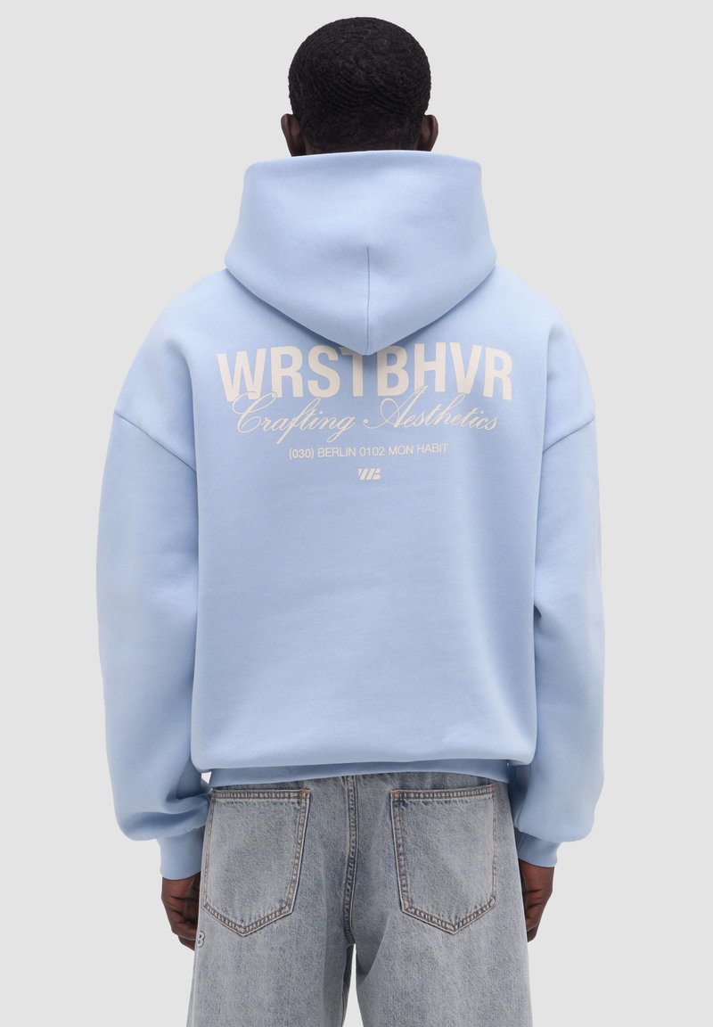 Mies, jolla on vaaleansininen huppari, jossa takaosassa teksti "WRSTBHVR Crafting Aesthetics (030) Berlin 0102 Mon Habit", yhdistettynä vaaleisiin denimhousuihin.