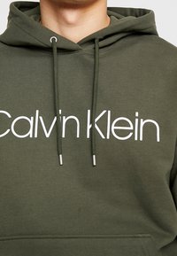 Felpa verde in cotone con un grande logo bianco "Calvin Klein" sul petto, dotata di lacci con estremità in argento e una finitura texture.